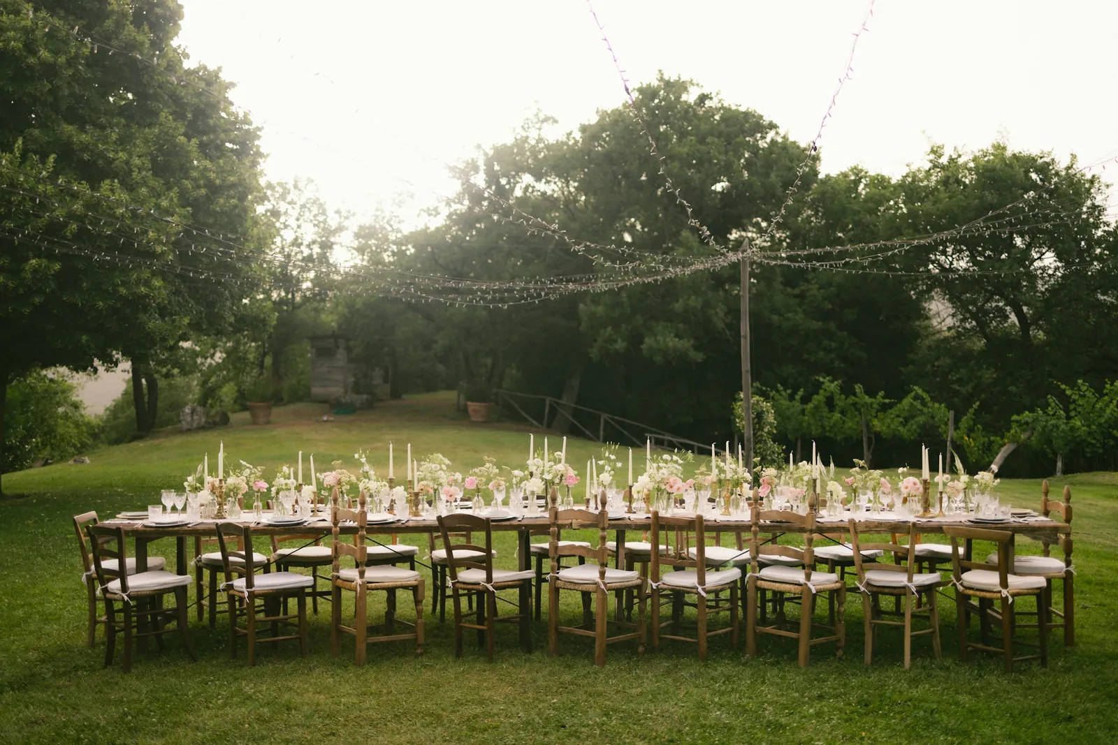 Long dinner table dressed on a garden lawn under trees and string lights at dusk