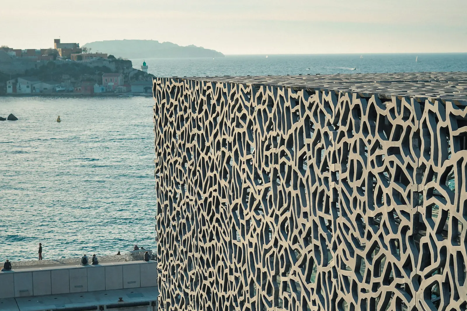 Latticed facade of the Mucem beside the sea in Marseille, with the harbour entrance and the coastline in late-afternoon light
