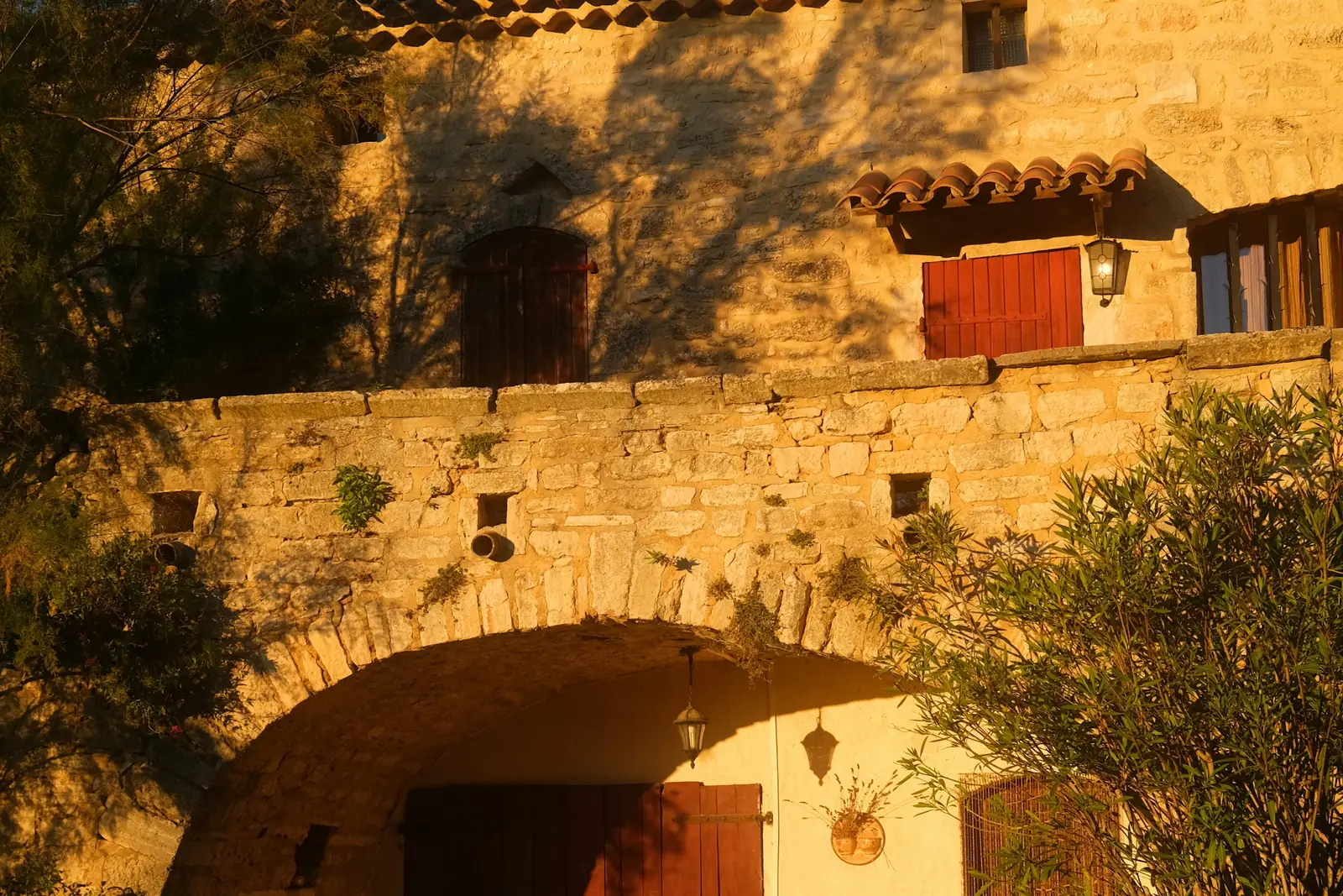 Old stone farmhouse with terracotta roof tiles, red shutters and an arched entrance in warm low sunlight